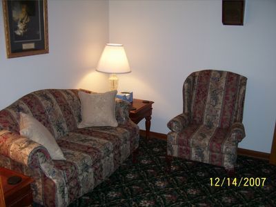 A corner of a room with a patterned sofa and armchair, a side table with a lamp, and a framed picture on the wall.