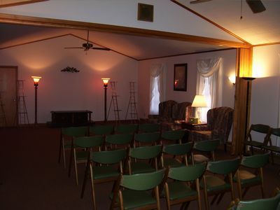 Rows of green chairs face a small stage in a dim, carpeted room with floor lamps and an armchair seating area.