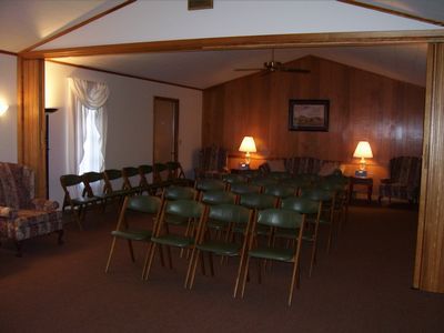 Rows of dark green folding chairs in a wood-paneled room with carpet, two lamps, and an armchair.