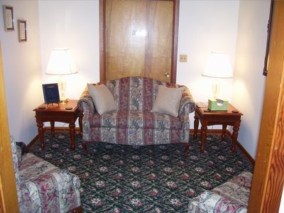 A small seating area with a striped loveseat flanked by two end tables, lamps, and armchairs on a patterned carpet.