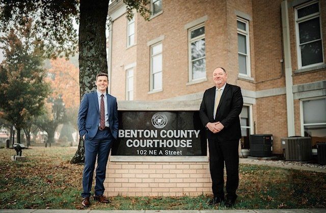 Two men are standing in front of a benton county courthouse sign.
