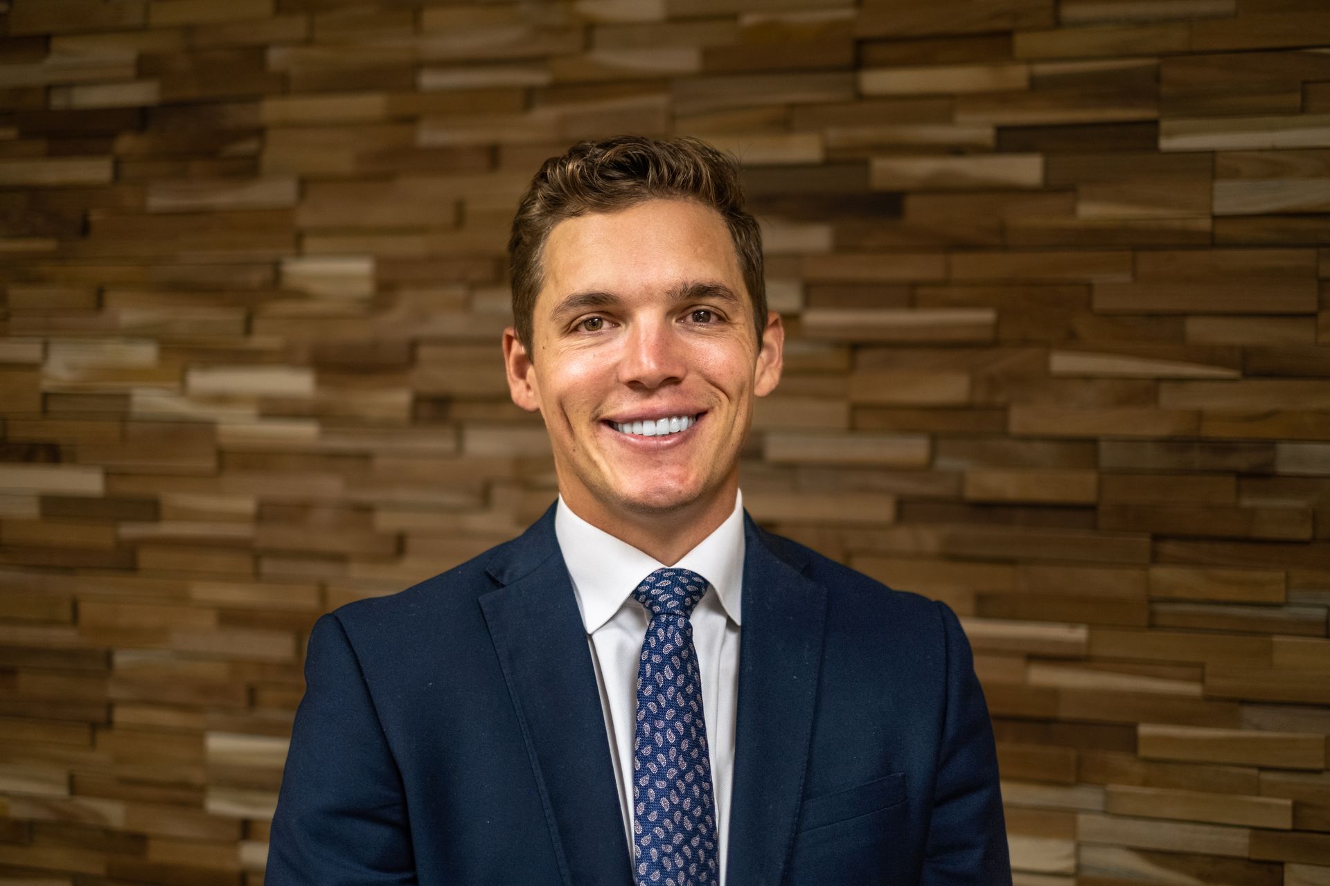 A man in a suit and tie is smiling in front of a wooden wall.