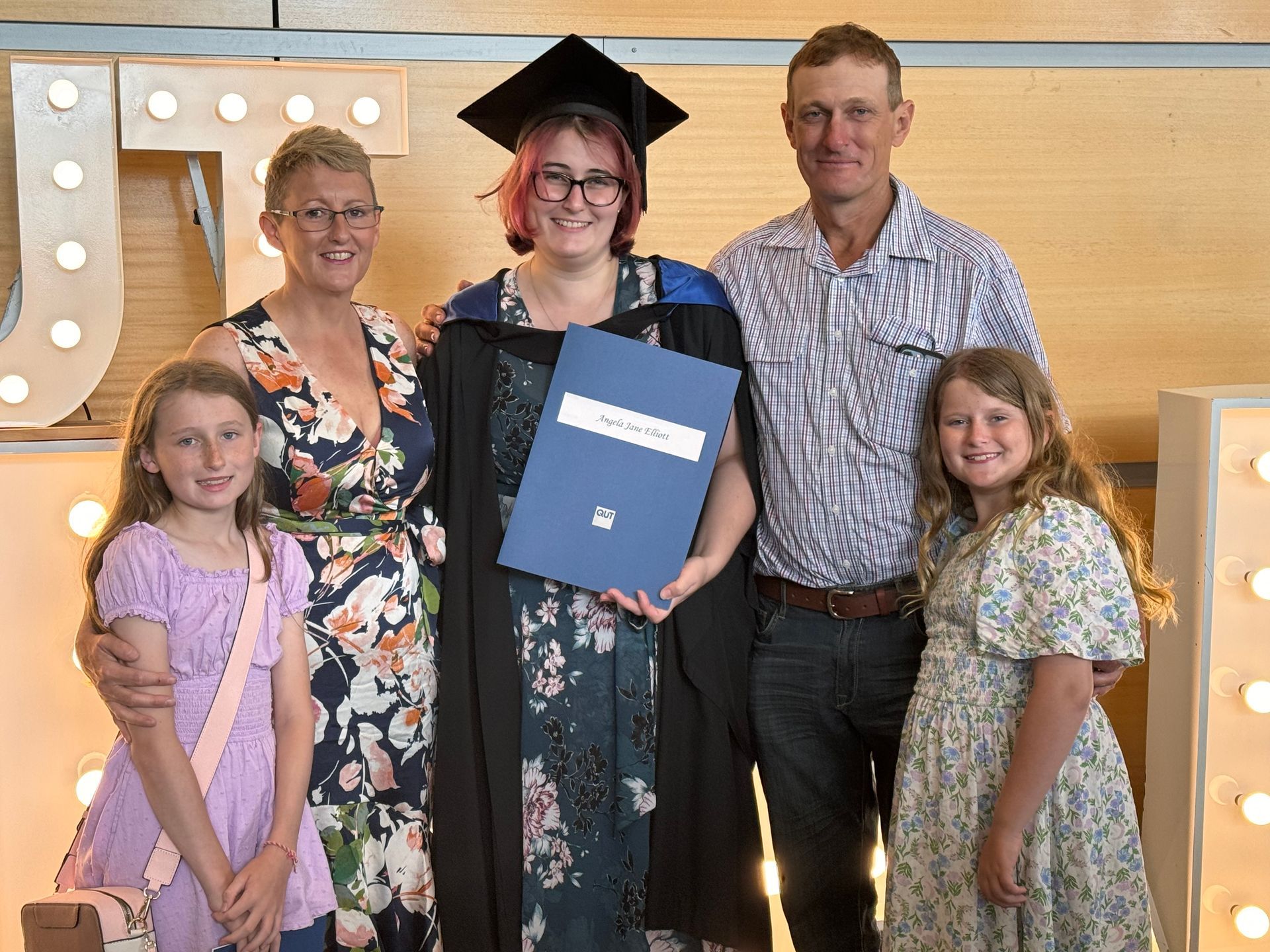 Family celebrates graduation: woman in cap and gown holds diploma, smiles with family in front of lit letters. — Gin Gin Domestic Water Supplies in Wallaville, QLD