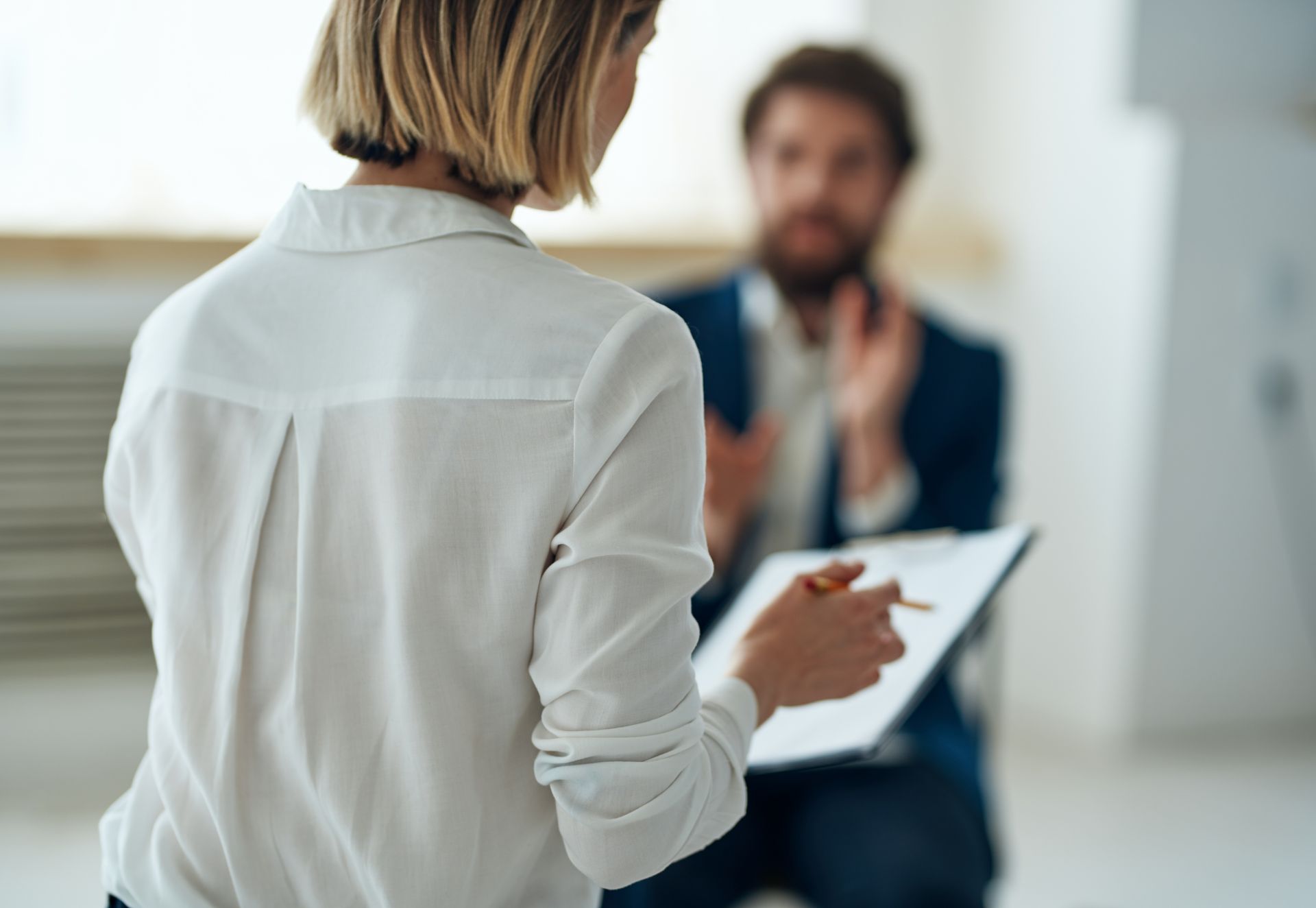 Woman taking notes during a conversation with a seated man in a bright office setting