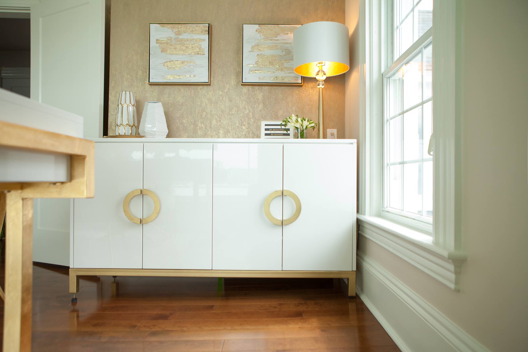 White cabinet with gold hardware, lamp, art, and window in a room with wood floors.