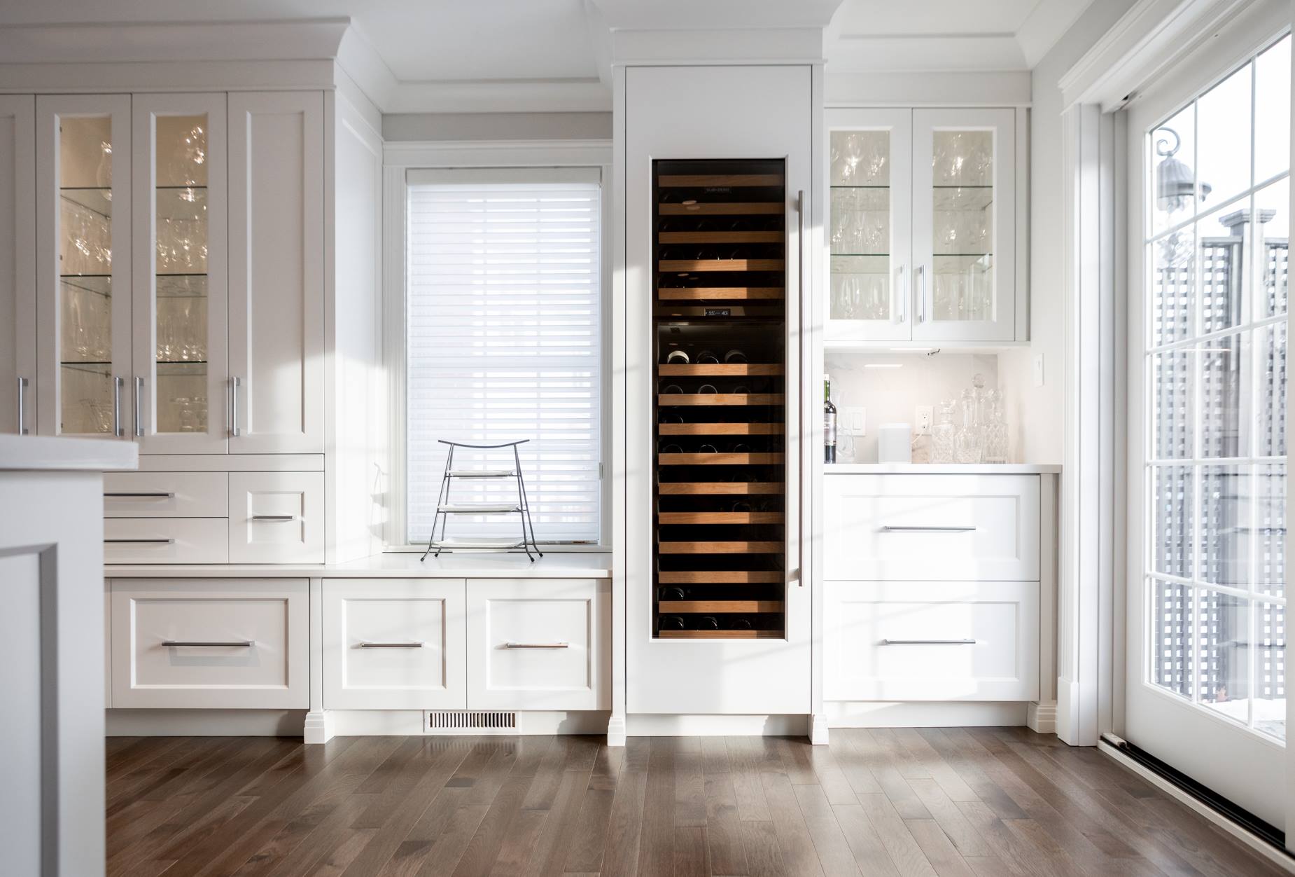 White kitchen with wine fridge, cabinets, and a window with blinds. Wooden floor and sunny light.
