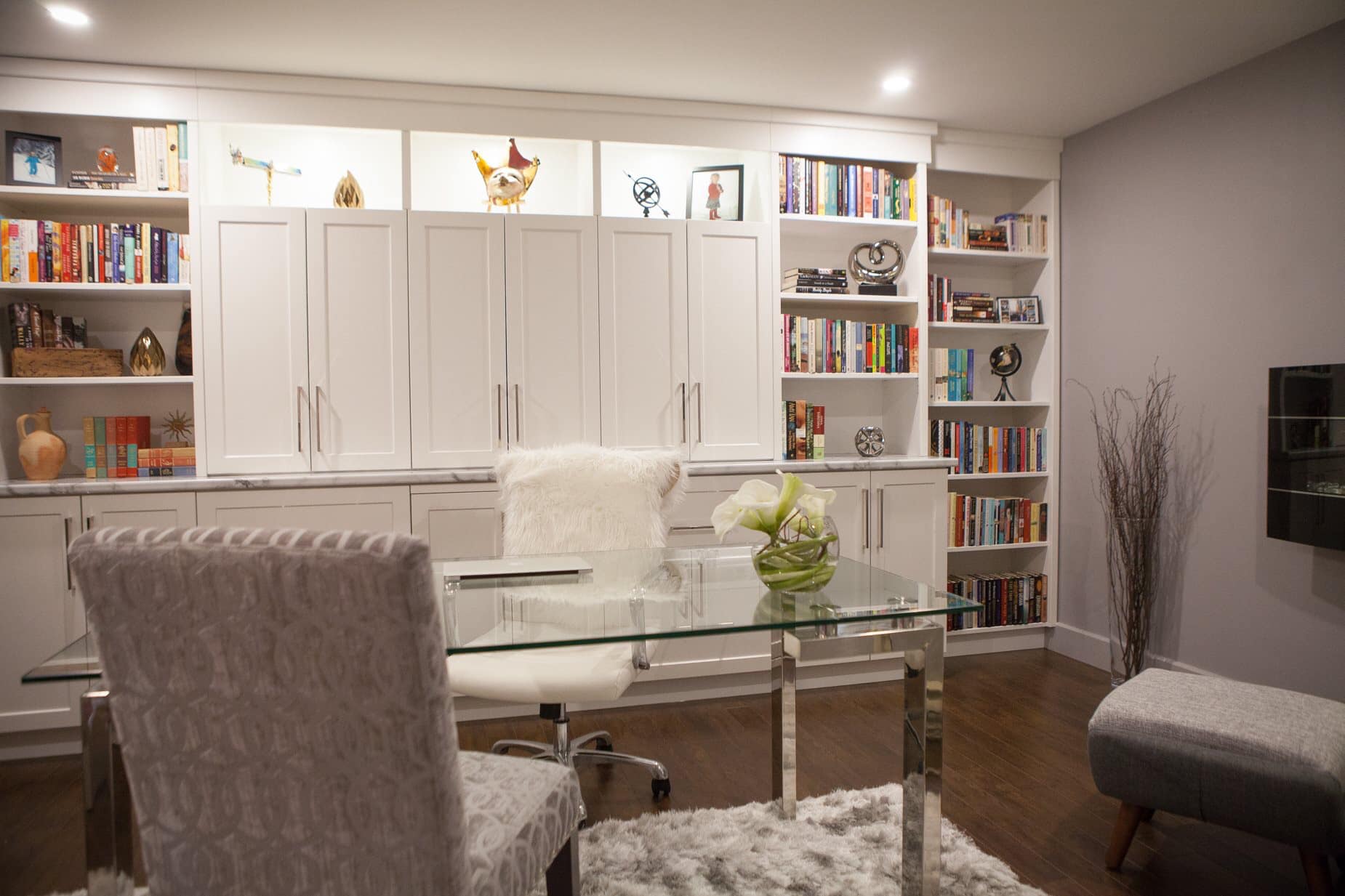 A modern home office with white built-in cabinets and shelves, glass desk, and fur chair.