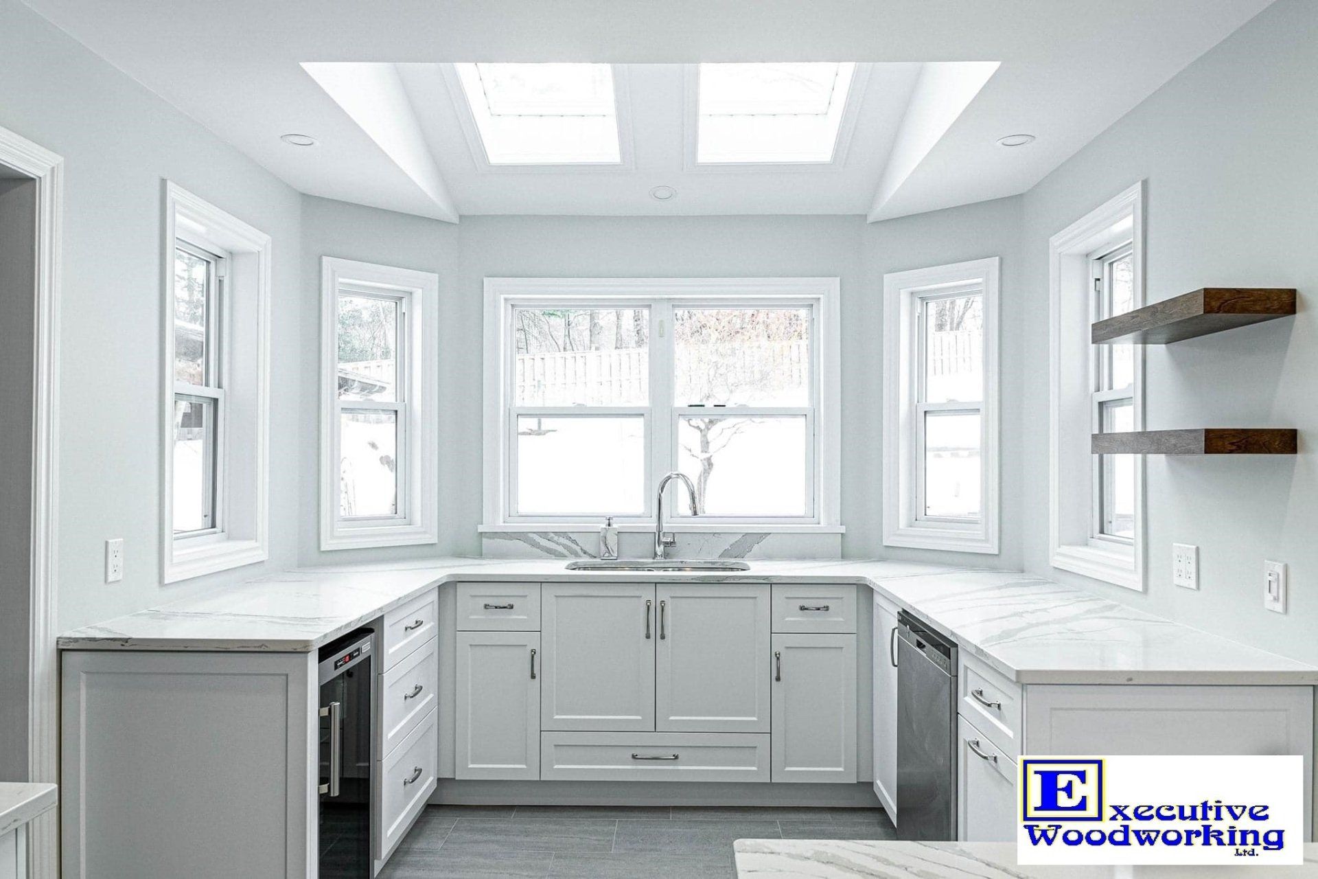 White U-shaped kitchen with marble countertops, windows, skylights, and wooden shelves.