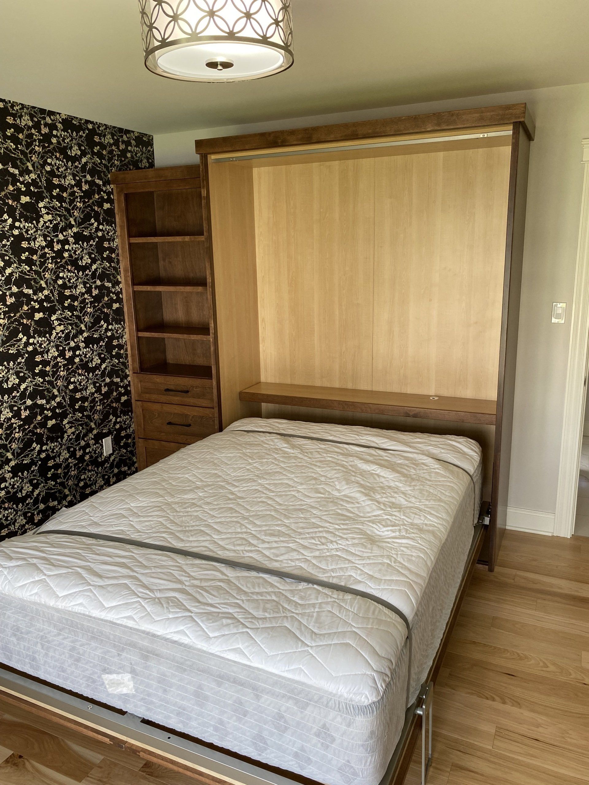 A wooden wall bed with a mattress, bookcase, and floral wallpaper.