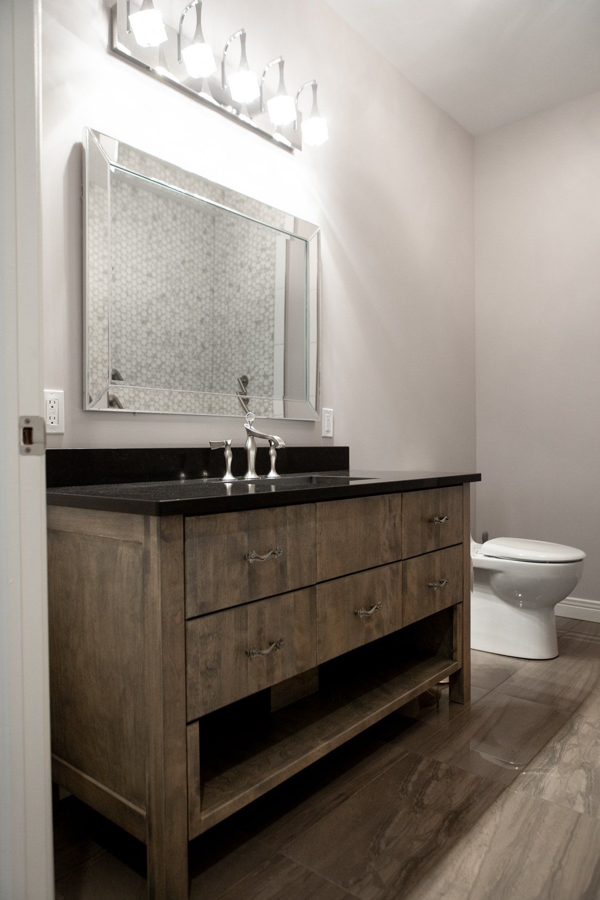 Bathroom with wooden vanity, black countertop, silver mirror, and white toilet.