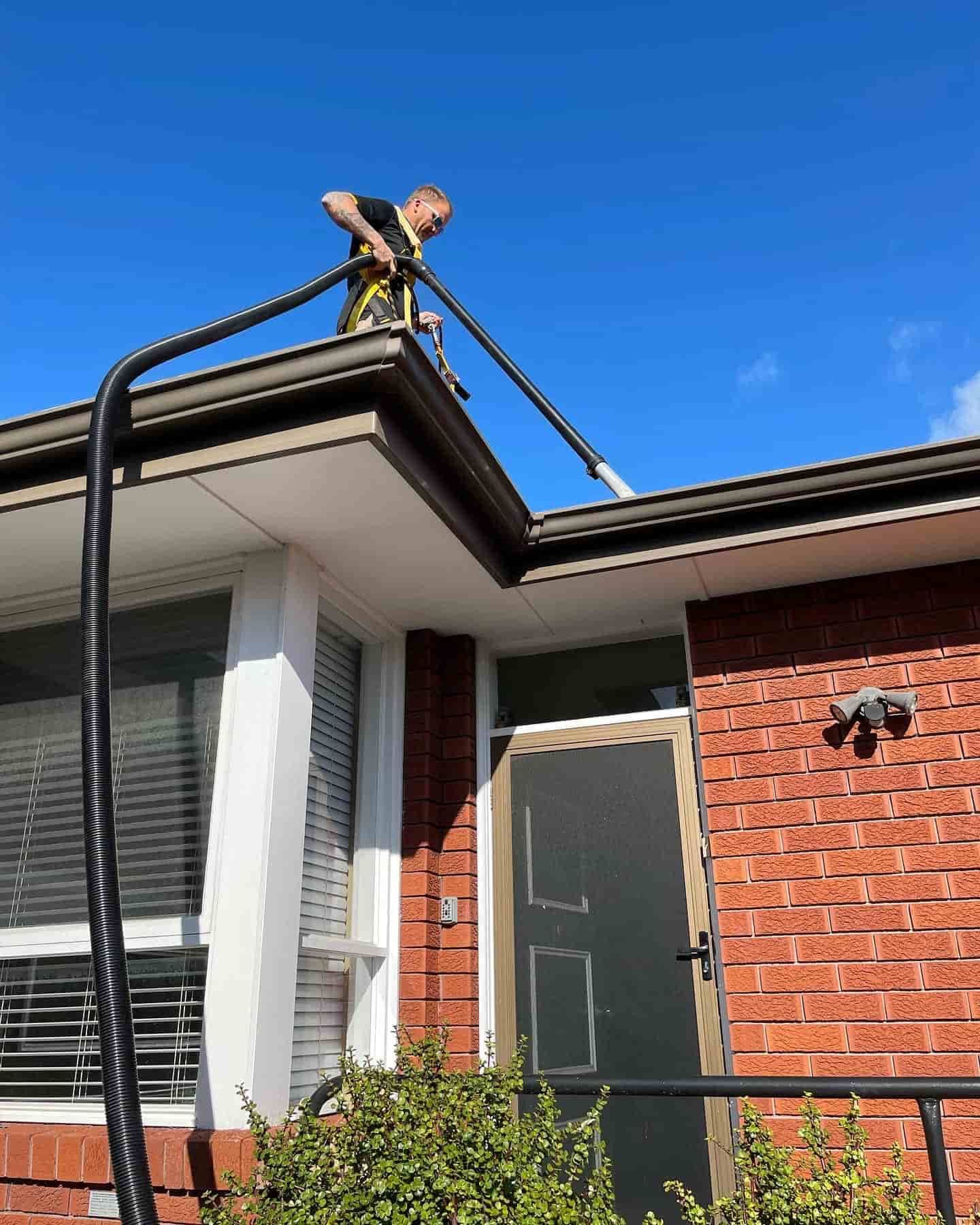 A Person on a Brick House Roof Uses a Long Hose to Vacuum Out the Gutters — South Coast Pressure Washer in Gerringong, NSW