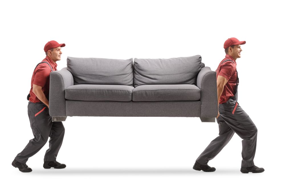 Two men are carrying a couch on their backs.