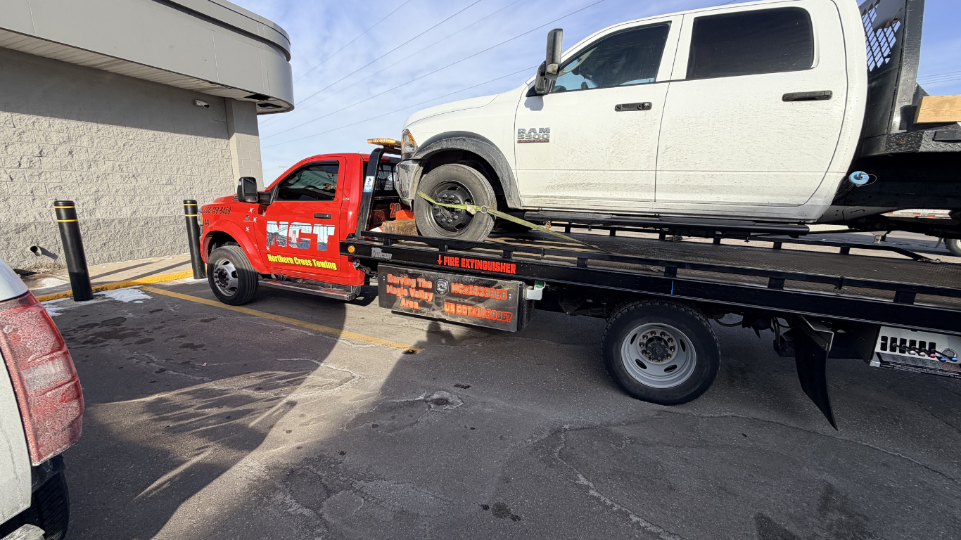 A bright red flatbed tow truck is hauling a white pickup truck in a parking lot.