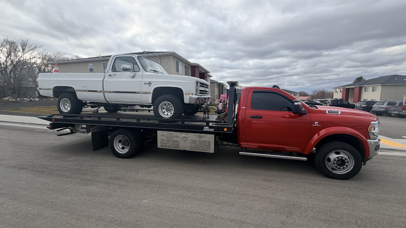 A red tow truck carrying a white vintage pickup truck on its flatbed in a paved lot.
