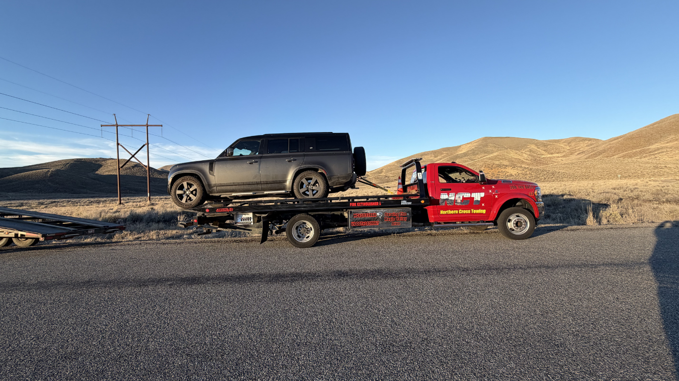 A red flatbed tow truck carries a dark gray SUV on a desert road with hills in the background under a blue sky.