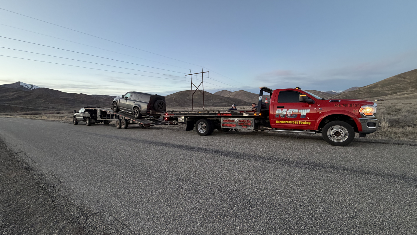 A red tow truck tows a trailer carrying a dark SUV along a paved road under a mountainous landscape at dusk.