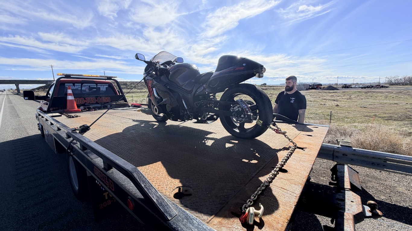 A person sits next to a black motorcycle secured on the flatbed of a tow truck on a sunny, open road.