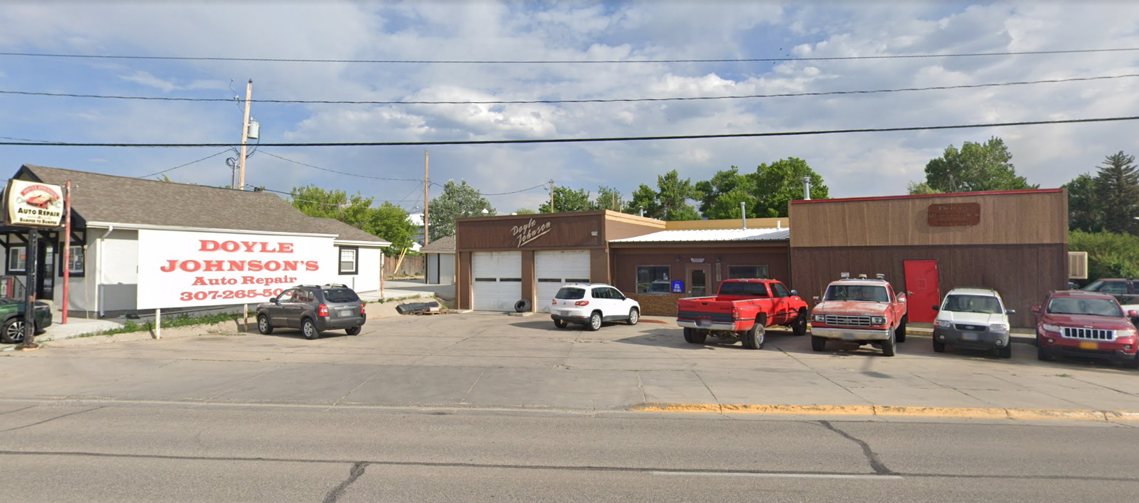Street view of a small brick auto shop with parked cars, a red pickup, and a white sign on the left