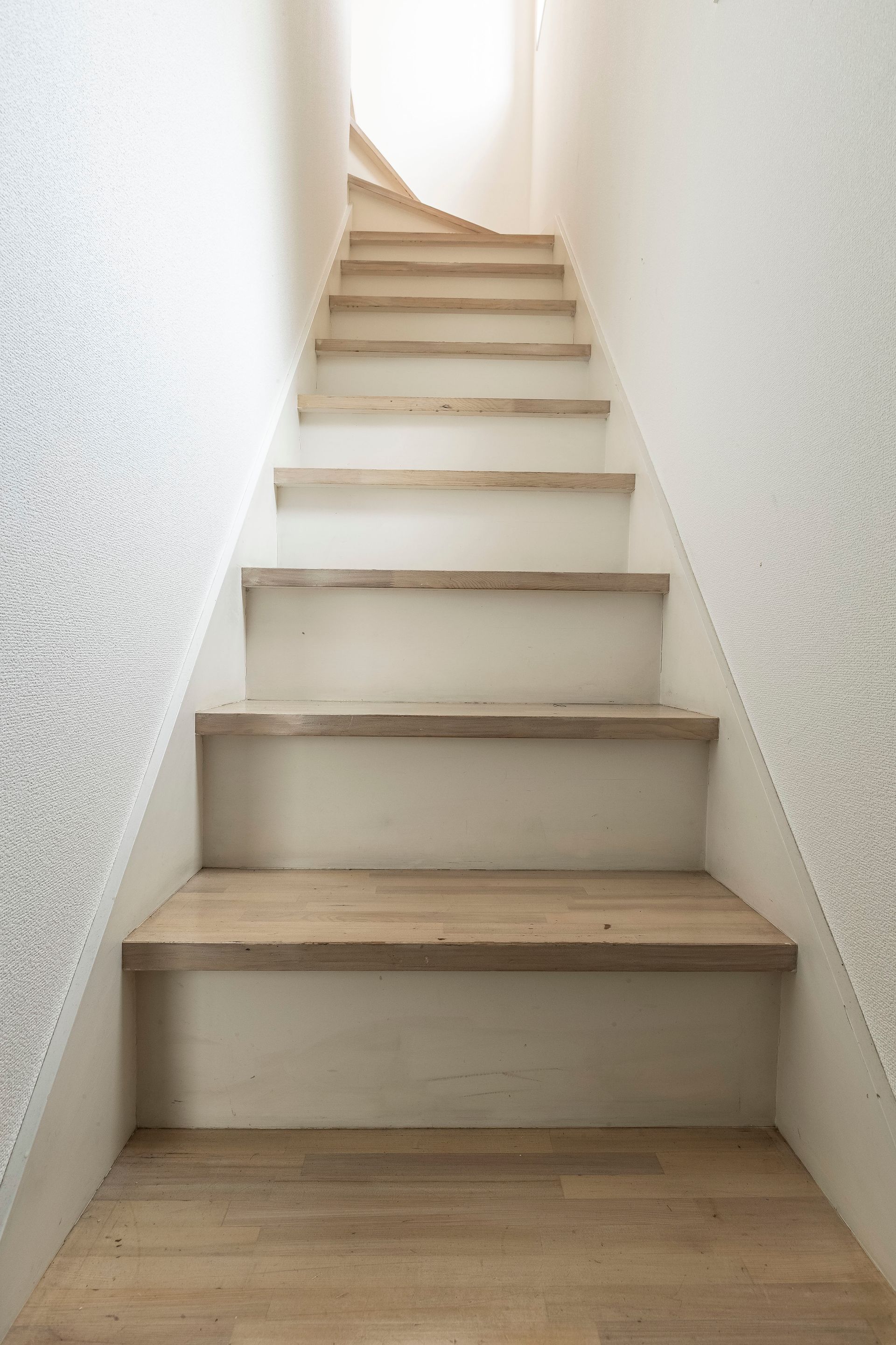 Staircase ascends between white walls with textured plaster and wood-toned steps, light from above.