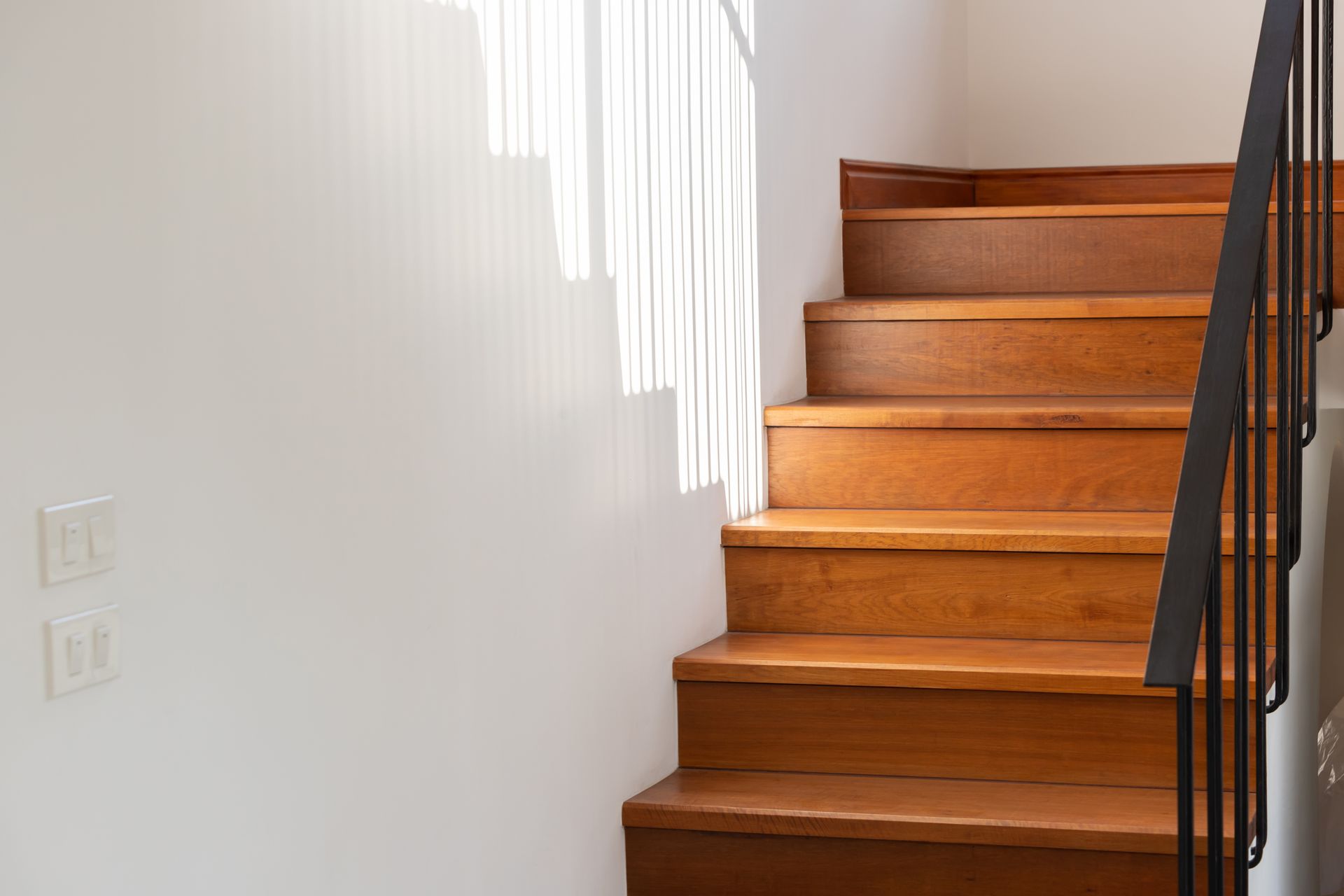 Wooden staircase with black metal railing, casting shadows on white wall.