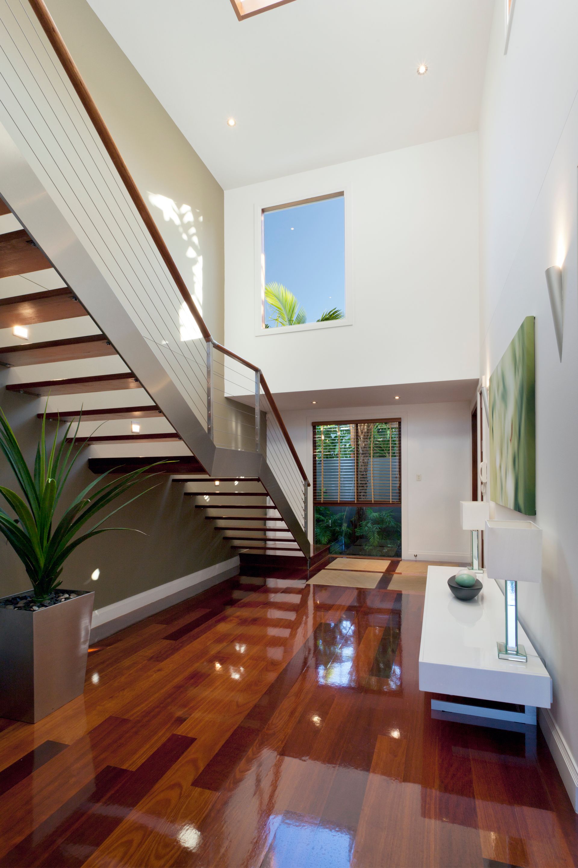 Modern home entryway with wooden floors, floating staircase, and large window overlooking trees.