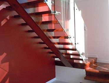Wooden staircase with glass railing. Red wall on the left, sunlight casts shadows.
