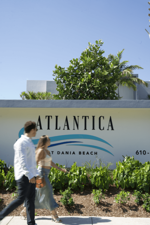 Couple walking past ATLANTICA sign and lush greenery under a clear blue sky at Atlantica at Dania Beach apartments in Dania Beach, FL.