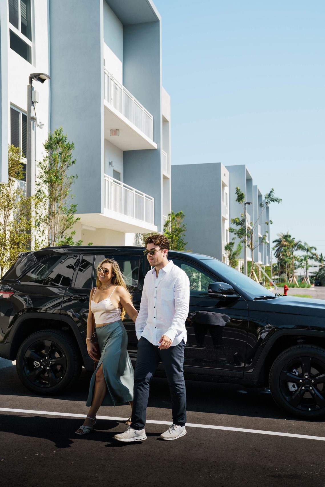 Two people standing beside a black SUV on a sunny street outside modern white buildings at Atlantica at Dania Beach apartments in Dania Beach, FL.