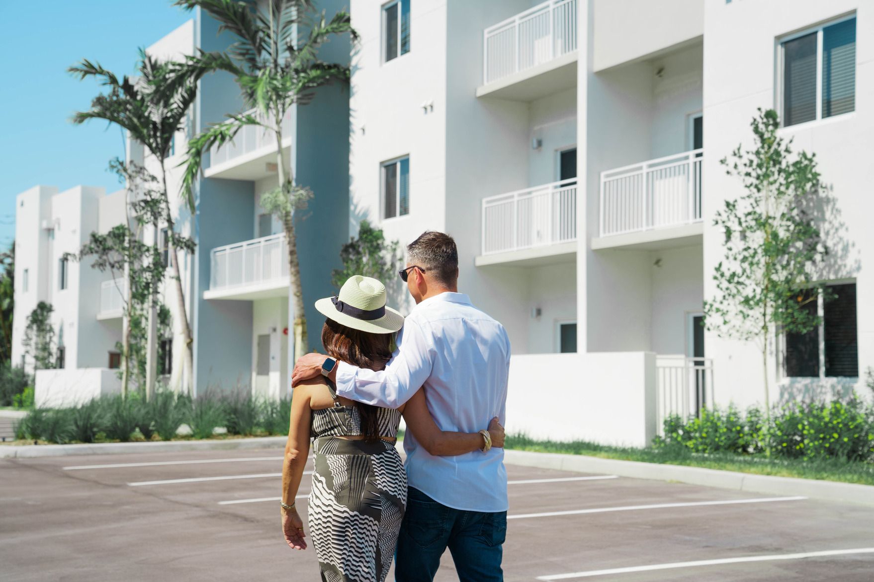 A couple walking arm-in-arm in a sunlit apartment parking lot beside white balconies and trees at Atlantica at Dania Beach apartments in Dania Beach, FL.