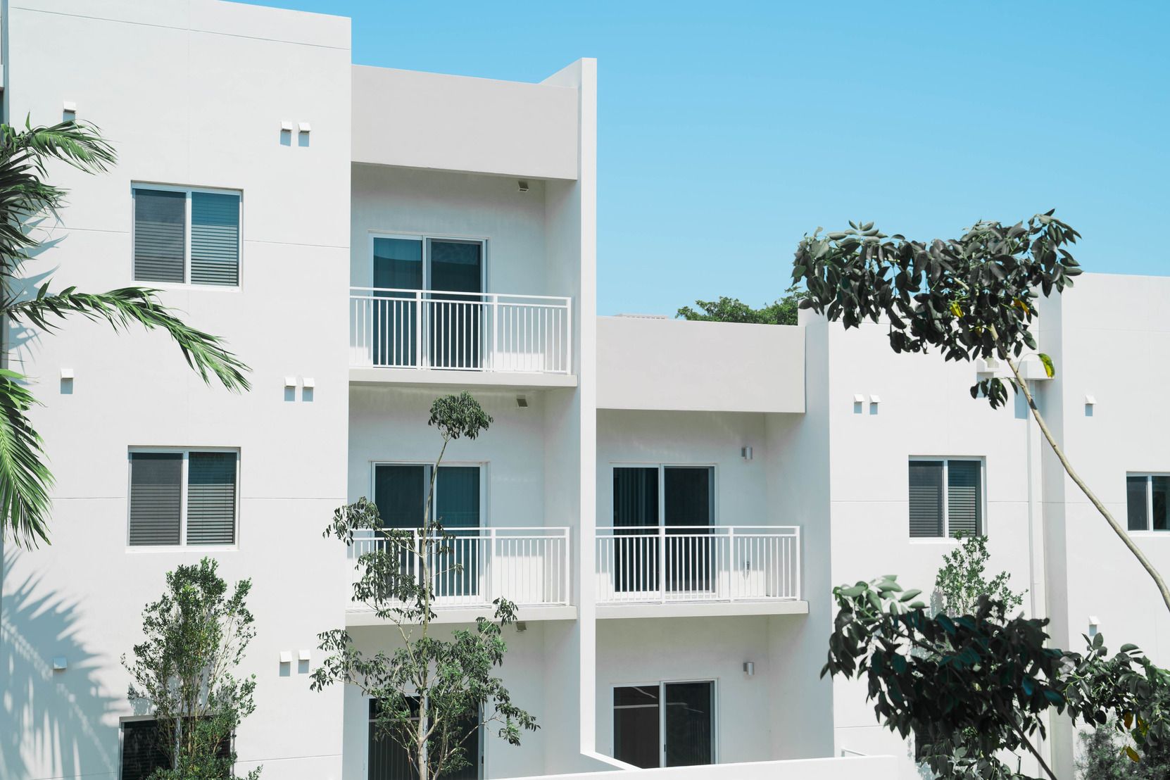 White apartment building with balconies and windows, framed by trees against a blue sky at Atlantica at Dania Beach apartments in Dania Beach, FL.