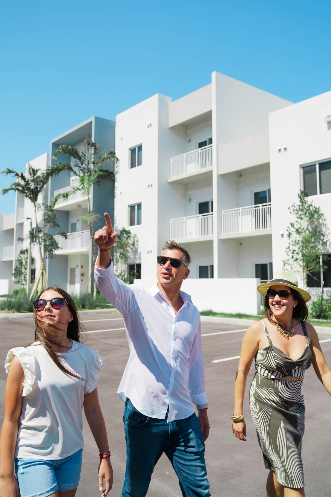 Three people walking outdoors in front of white apartment buildings, smiling under a blue sky at Atlantica at Dania Beach apartments in Dania Beach, FL.