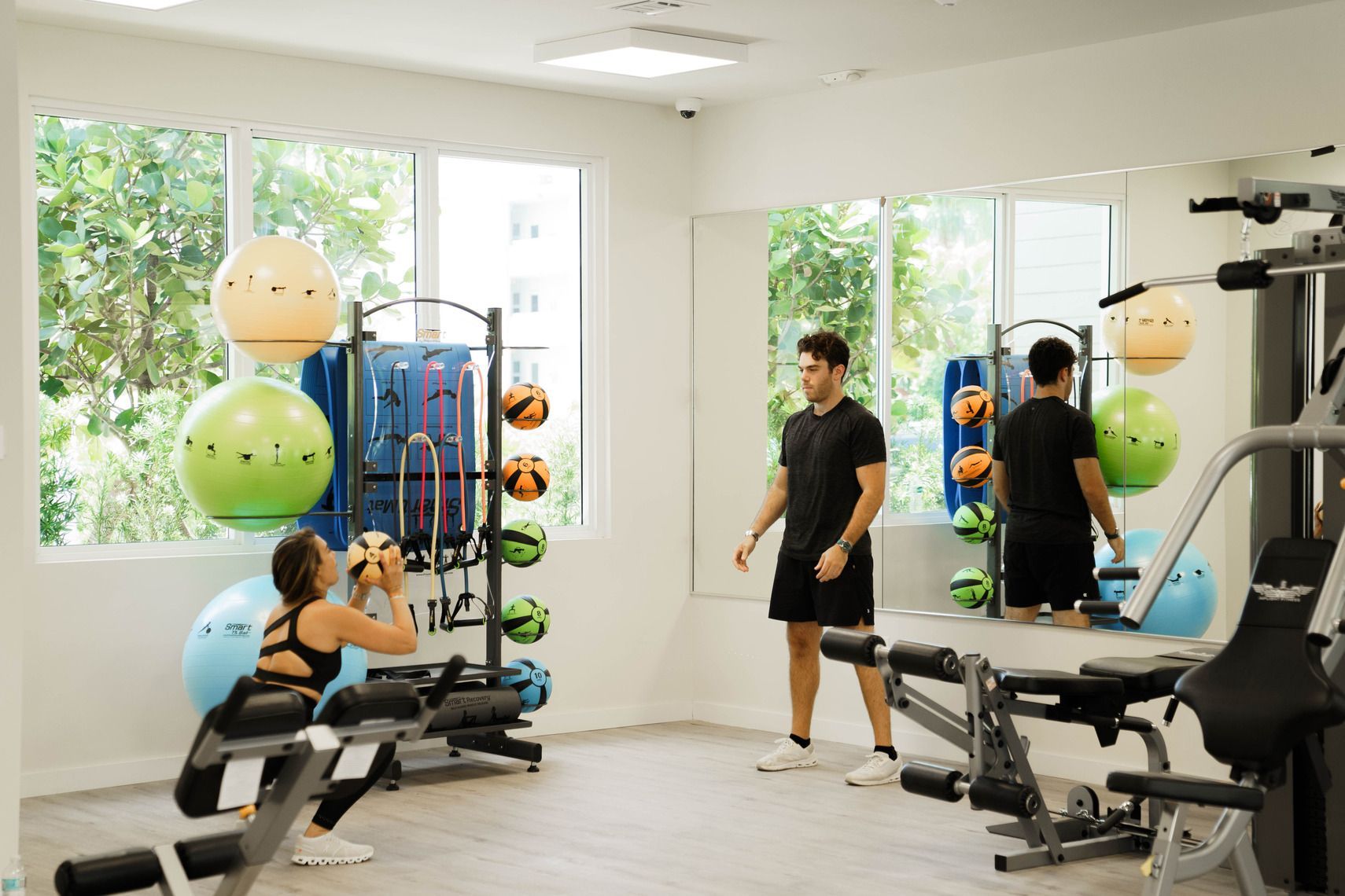 Bright gym with people exercising on machines, medicine balls, and weights near large windows at Atlantica at Dania Beach apartments in Dania Beach, FL.