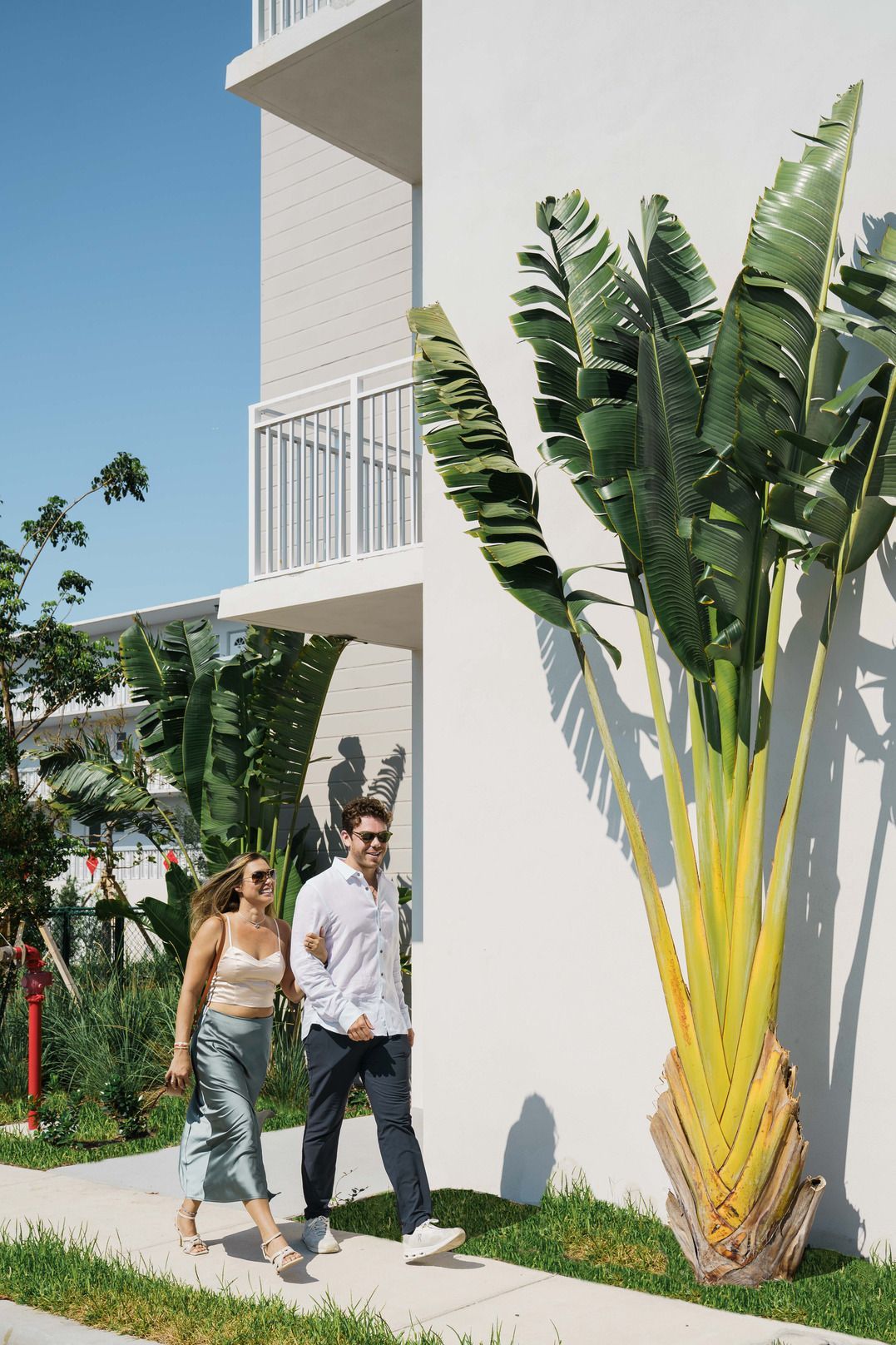 Two people walking beside a white building with a tall banana plant and blue sky at Atlantica at Dania Beach apartments in Dania Beach, FL.