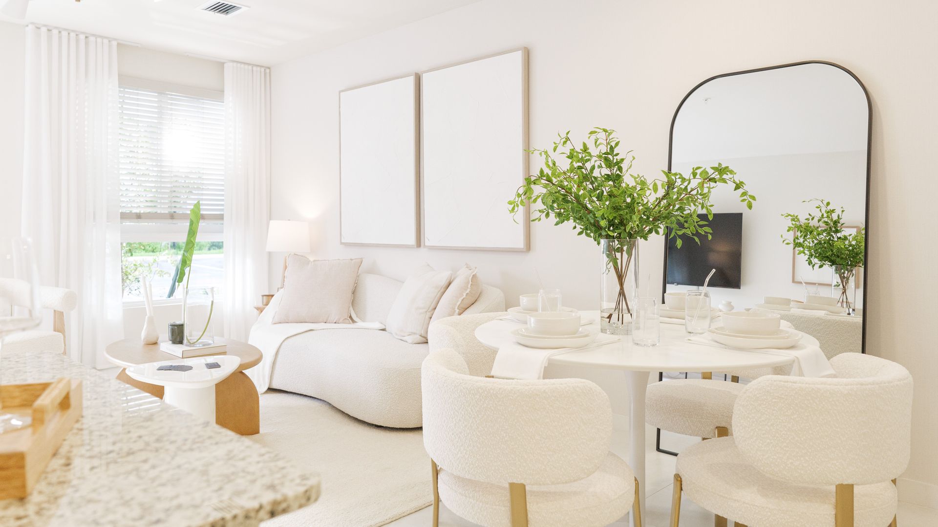 Bright white modern living room with a round dining table, cream chairs, large mirror, and plants at Atlantica at Dania Beach apartments in Dania Beach, FL.