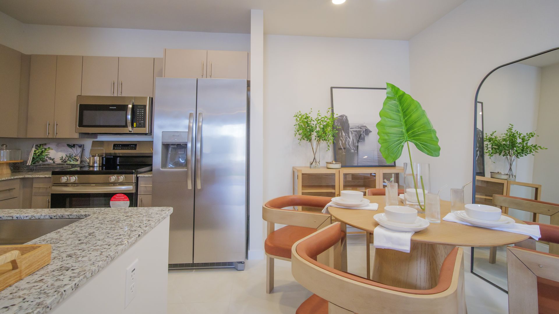 Modern kitchen and dining area with granite island, stainless fridge, and round table by a large plant-shelved window at Atlantica at Dania Beach apartments in Dania Beach, FL.