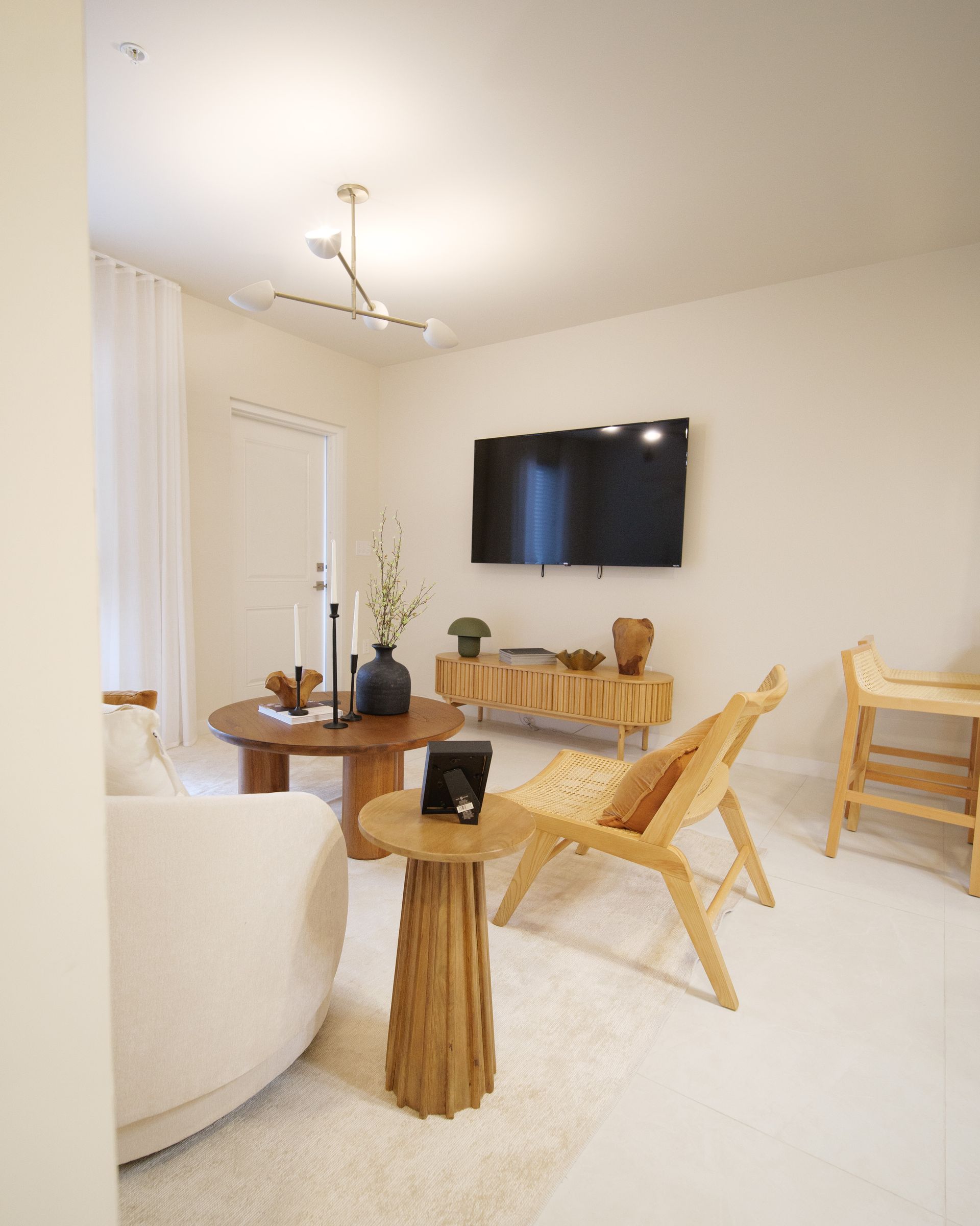 Bright minimalist living room with wall-mounted TV, round wooden tables, wicker chair, and small dining set at Atlantica at Dania Beach apartments in Dania Beach, FL.
