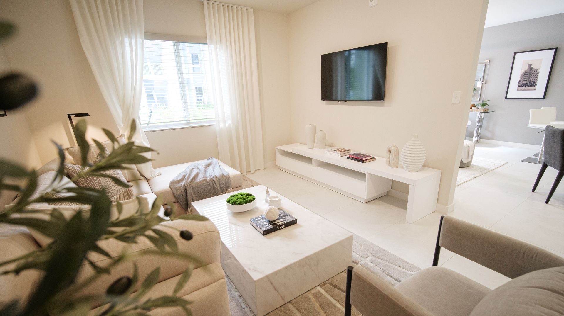 Bright beige living room with white furniture, wall-mounted TV, coffee table, and window with blinds at Atlantica at Dania Beach apartments in Dania Beach, FL.
