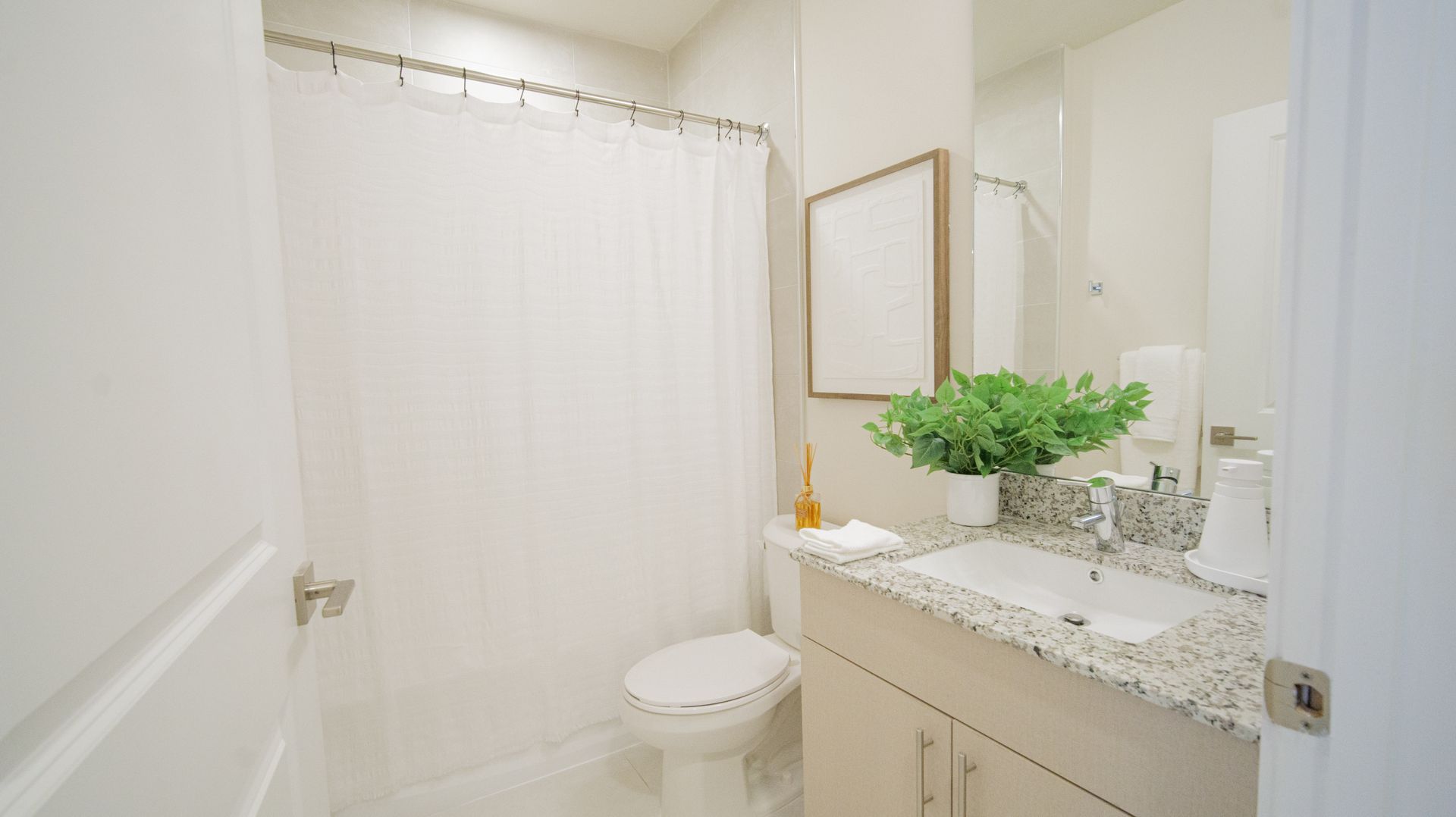 Bright white bathroom with shower curtain, sink vanity, mirror, and potted greenery at Atlantica at Dania Beach apartments in Dania Beach, FL.