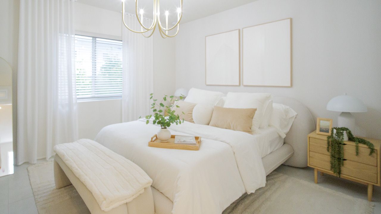 Bright white bedroom with a bed, bench, chandelier, window shutters, and a wicker basket on the floor at Atlantica at Dania Beach apartments in Dania Beach, FL.