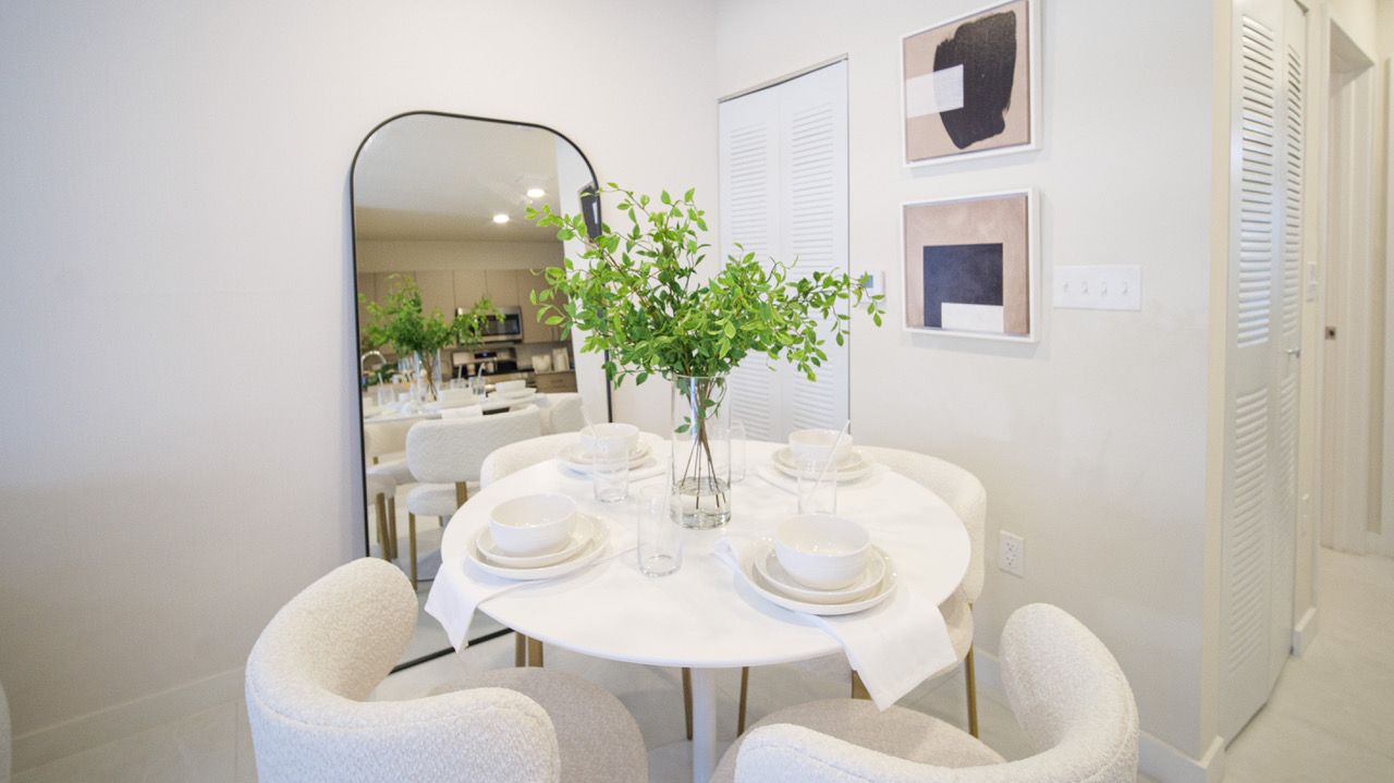 Bright dining nook with white table, upholstered chairs, mirror, and green flowers in a minimalist room at Atlantica at Dania Beach apartments in Dania Beach, FL.