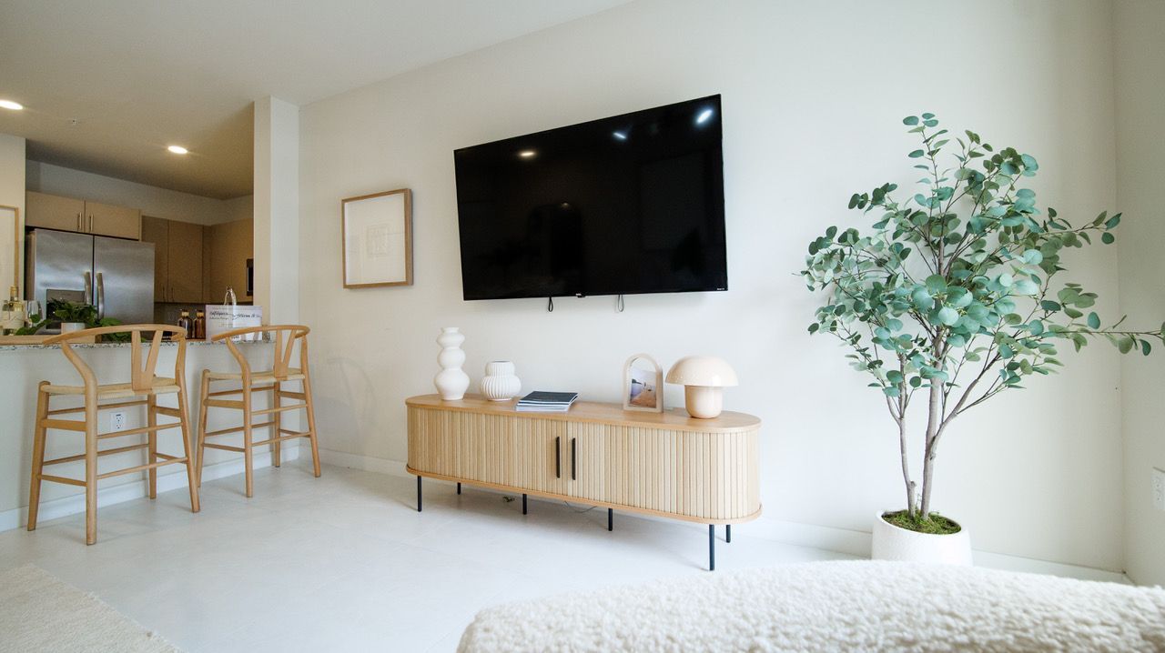 Bright minimalist living room with wall-mounted TV, wood console, bar stools, and potted tree by a rug at Atlantica at Dania Beach apartments in Dania Beach, FL.