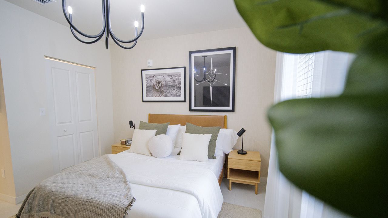 Bright bedroom with a white bed, tan headboard, bedside table, framed wall art, and large window curtains at Atlantica at Dania Beach apartments in Dania Beach, FL.