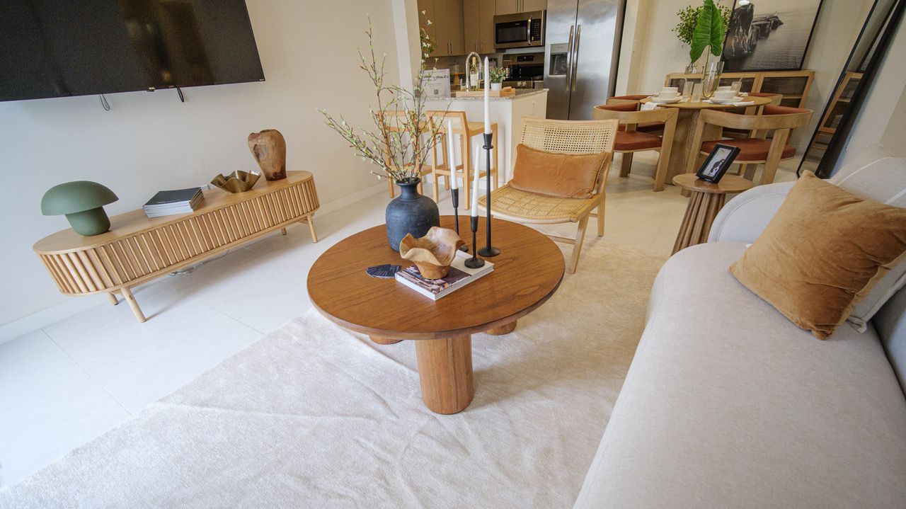 Bright living room with round wooden coffee table, beige sofa, wicker chair, and neutral decor at Atlantica at Dania Beach apartments in Dania Beach, FL.