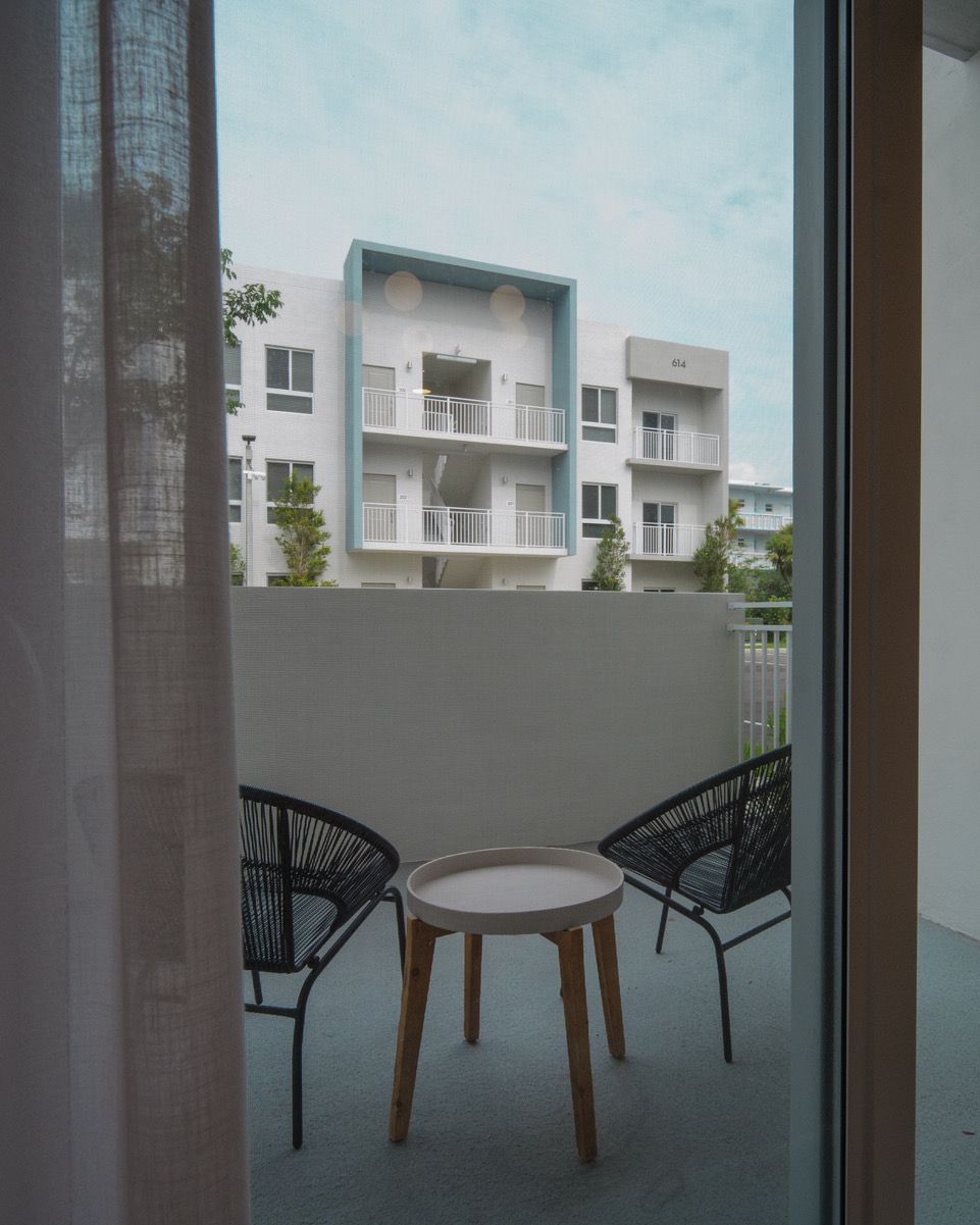 Balcony with two black chairs and a small round table overlooking modern apartment buildings at Atlantica at Dania Beach apartments in Dania Beach, FL.