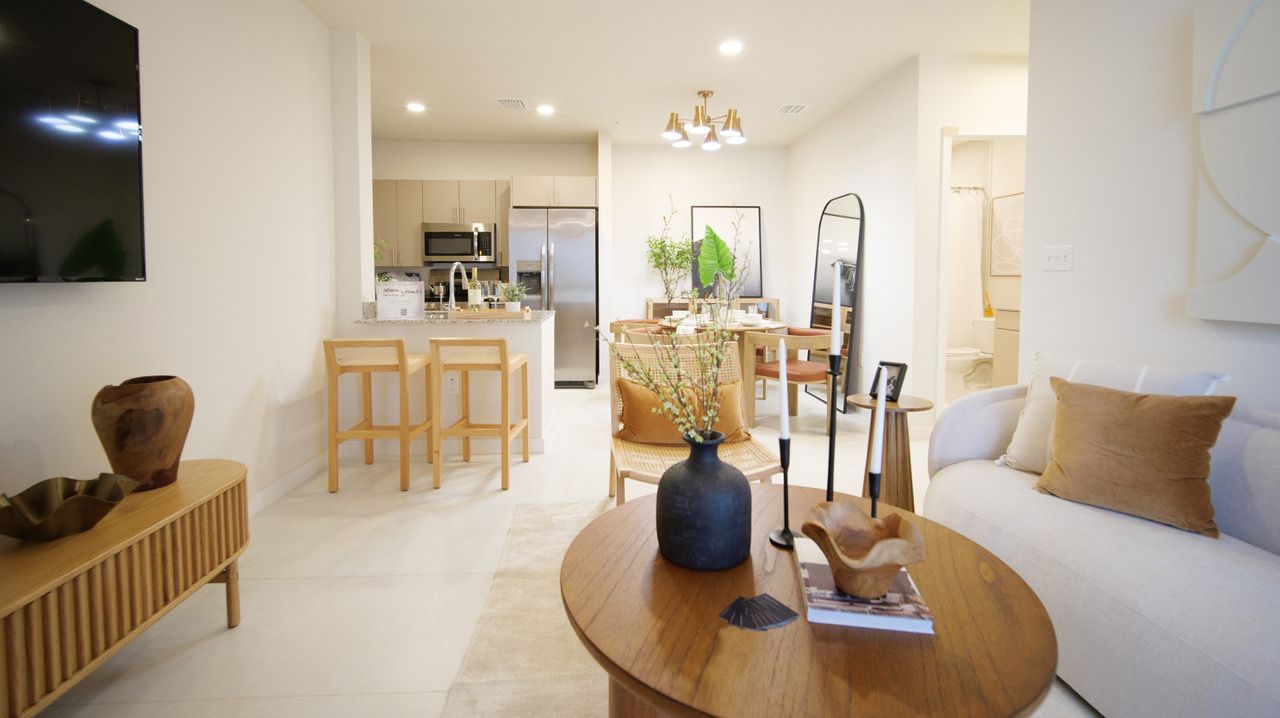 Bright open-plan living room with wood table, beige sofa, bar stools, and kitchen in the background at Atlantica at Dania Beach apartments in Dania Beach, FL.