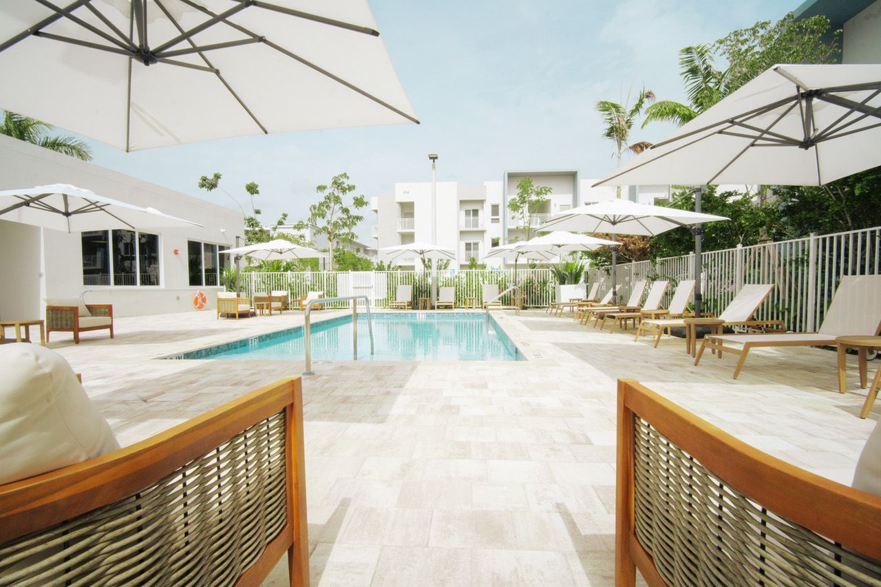 Sunny poolside courtyard with lounge chairs, white umbrellas, and modern buildings around a turquoise pool at Atlantica at Dania Beach apartments in Dania Beach, FL.