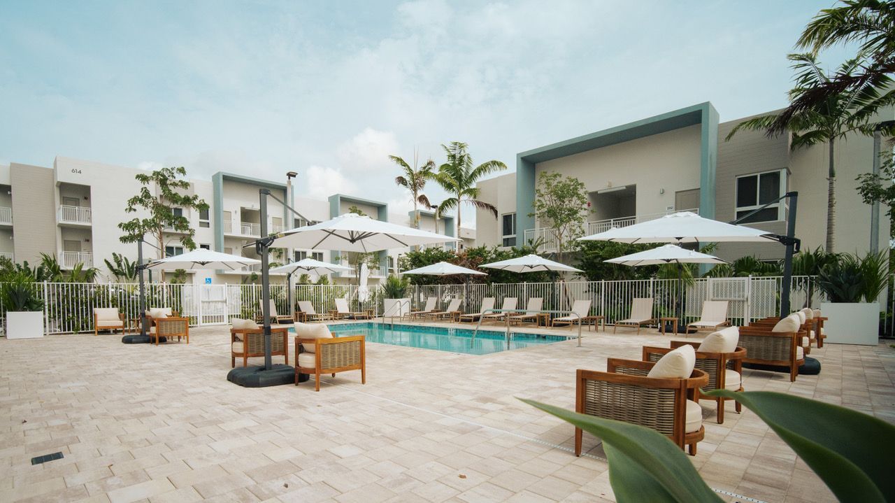 Resort pool deck with lounge chairs, white umbrellas, and modern buildings in a sunny courtyard at Atlantica at Dania Beach apartments in Dania Beach, FL.