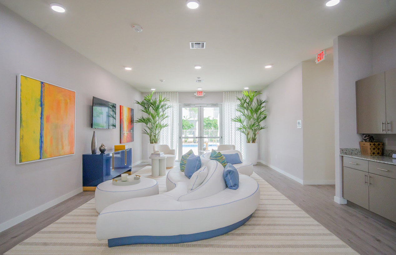Bright modern lounge with white curved seating, blue pillows, colorful wall art, and large windows at Atlantica at Dania Beach apartments in Dania Beach, FL.