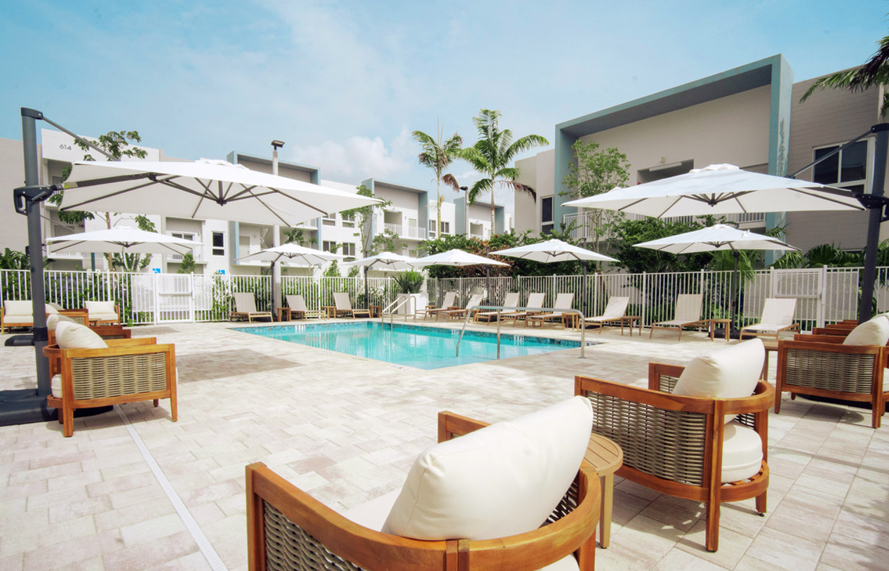 Modern resort pool area with lounge chairs, umbrellas, and white buildings under a bright sky at Atlantica at Dania Beach apartments in Dania Beach, FL.