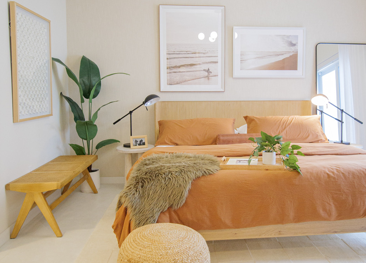Bright minimalist bedroom with an orange bedspread, wooden bench, plants, and framed wall art at Atlantica at Dania Beach apartments in Dania Beach, FL.